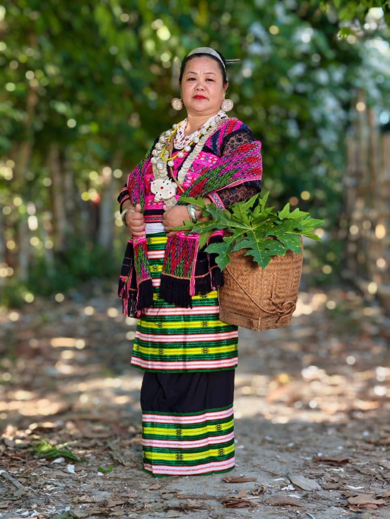 A Mishmi woman in traditional woven attire and silver jewellery, carrying a woven basket of greens.