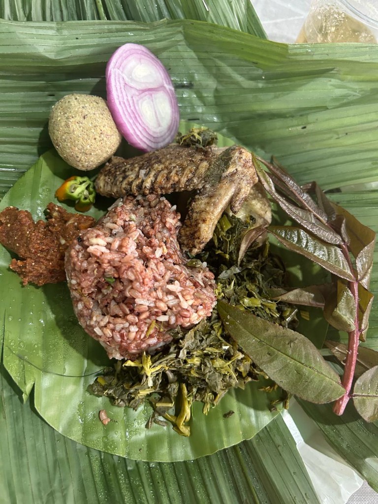 A traditional meal with local rice, greens, and sides served on a large green leaf.